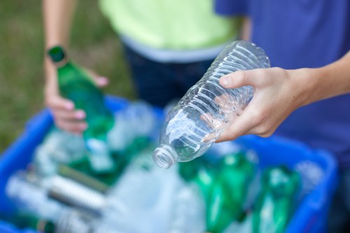 A close-up view of a person's hand holding a clear plastic water bottle, with the bottle slightly tilted and empty of liquid. In the background, there are two other individuals, one in a yellow top and the other in blue, with only their torsos visible. The scene appears to take place outdoors on a grassy area, with a blue recycling bin filled with various green and transparent plastic bottles and other rubbish items. The environment suggests a waste management or rubbish clearance activity, consistent with services offered by Garden Clearance Brent Cross in the Brent Cross area, where rubbish disposal and recycling of plastics are common tasks. The focus on the plastic bottles and the outdoor setting highlights the importance of proper rubbish disposal and recycling efforts in the local community.