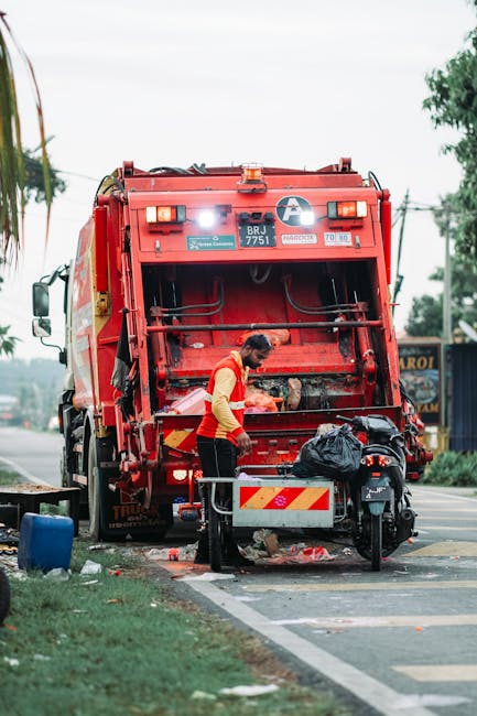 A commercial waste collection vehicle is parked on a narrow urban street, positioned parallel to a row of old, multi-story buildings with stone and brick facades. The vehicle's rear hatch is open, rev