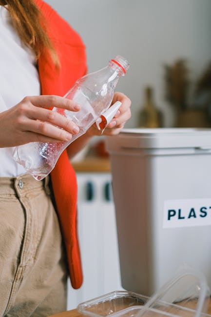 Close-up view of a person's gloved hand gripping the top of a large, semi-transparent blue plastic rubbish bag filled with waste. The individual is wearing an orange protective suit, and the bag appea