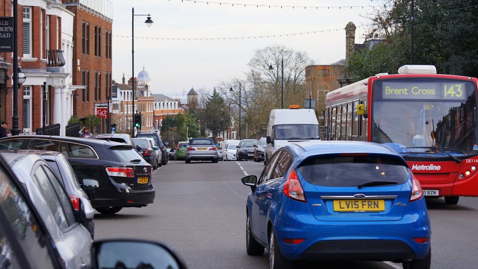 A busy urban street scene featuring parked cars lining both sides of the road, including a black vehicle and a blue hatchback in the foreground. In the midground, a red Metroline bus displaying route